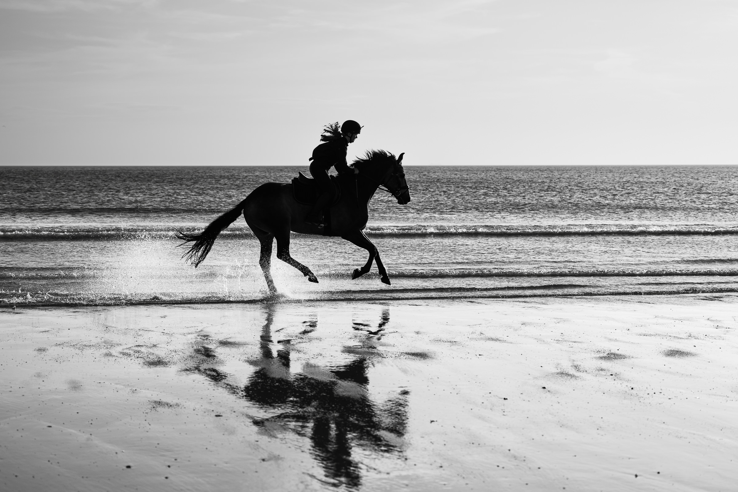 black and white photo of a horse and rider cantering on a beach