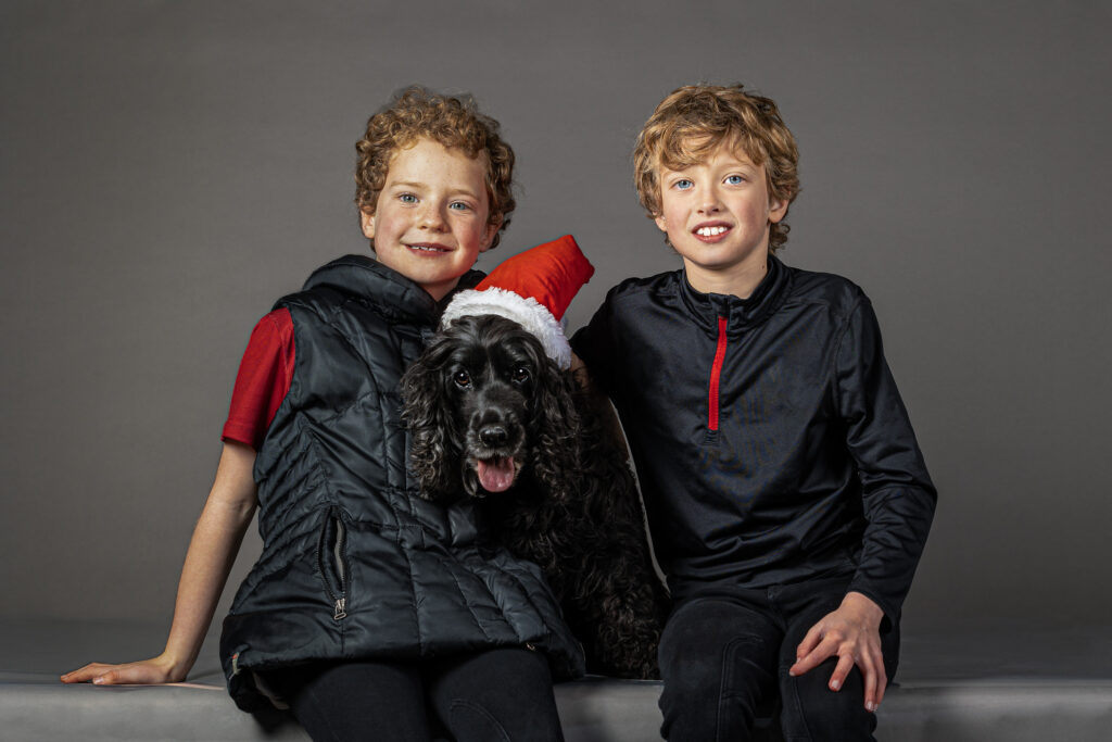 pet portrait photo with two boys and a black cocker spaniel with red Santa hat and a studio background