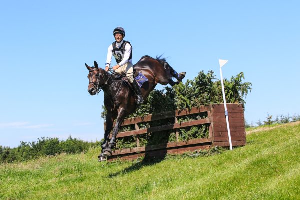Brown horse and rider jumping a cross country fence in competition with blue sky and green grass
